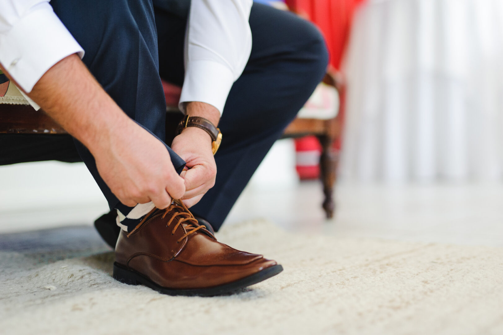 A person in formal attire sits on a chair, meticulously tying brown leather dress shoes, preparing for a refined ceremony at Congress Hall or a Cape May wedding.