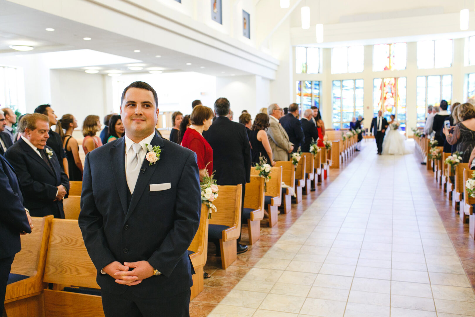 A man in a suit stands in a church aisle, reminiscent of Congress Hall's grandeur, with guests seated on either side. At the front, near a stained glass window, a couple exchanges vows, evoking the charm of a Cape May wedding.