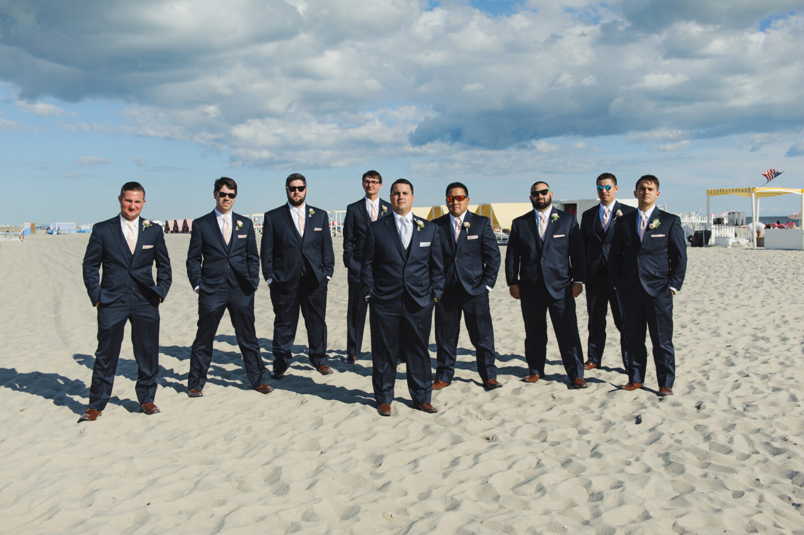 A group of nine men in suits stands on a sandy beach at Congress Hall, Cape May, with a cloudy sky providing the perfect backdrop for a stylish wedding photograph.