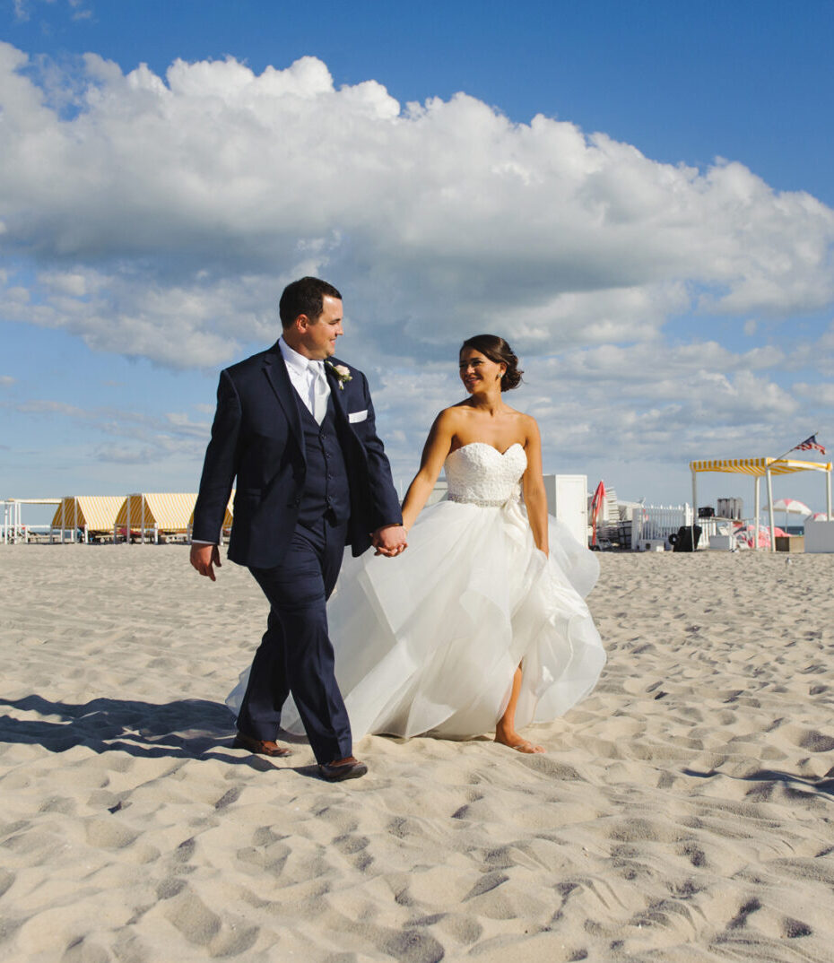 A couple in formal attire strolls barefoot along the sandy beach under a blue sky with clouds, their minds filled with memories of their enchanting Congress Hall Cape May wedding.