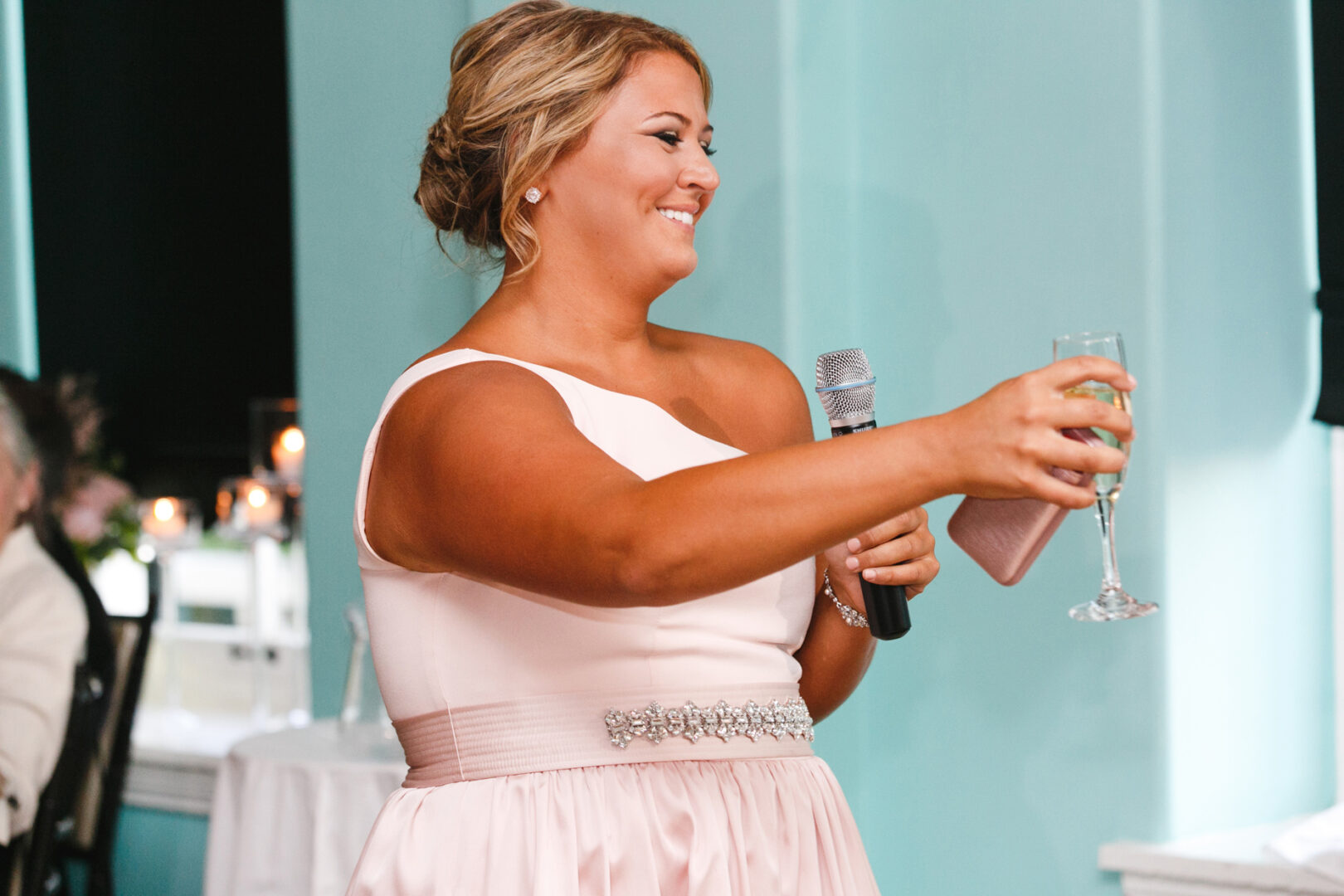 At a Congress Hall Cape May wedding, a woman in a pink dress holds a microphone and champagne glass, smiling warmly. She stands indoors against teal walls with candles flickering softly in the background.