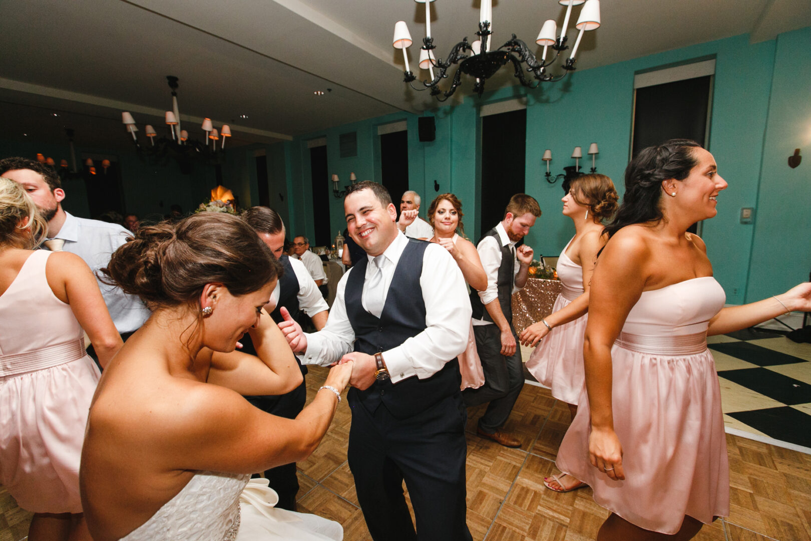At a charming indoor event at Congress Hall in Cape May, a group of people in formal attire gracefully dance beneath shimmering chandeliers against the backdrop of a turquoise wall.
