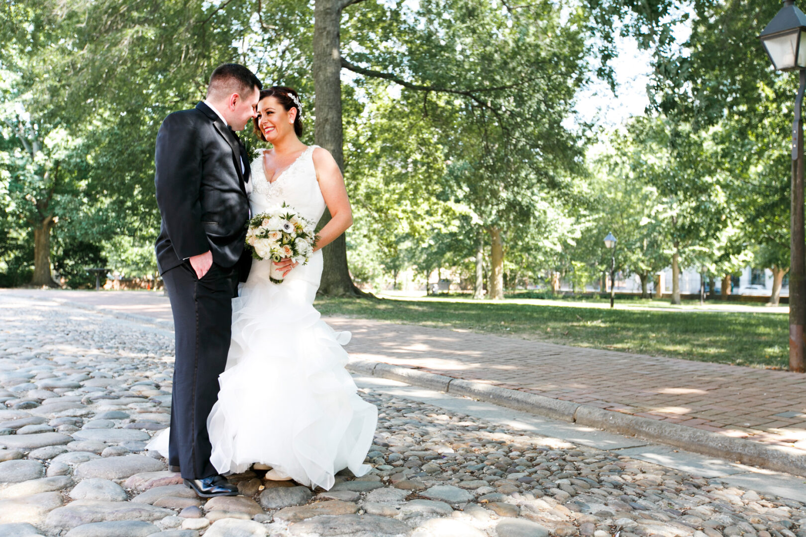 On a sunny day, a bride and groom stand closely on a cobblestone path beneath the trees, her white dress shimmering as she holds a bouquet. He's dapper in his suit. The scene captures the elegance of their Ballroom at the Ben wedding.