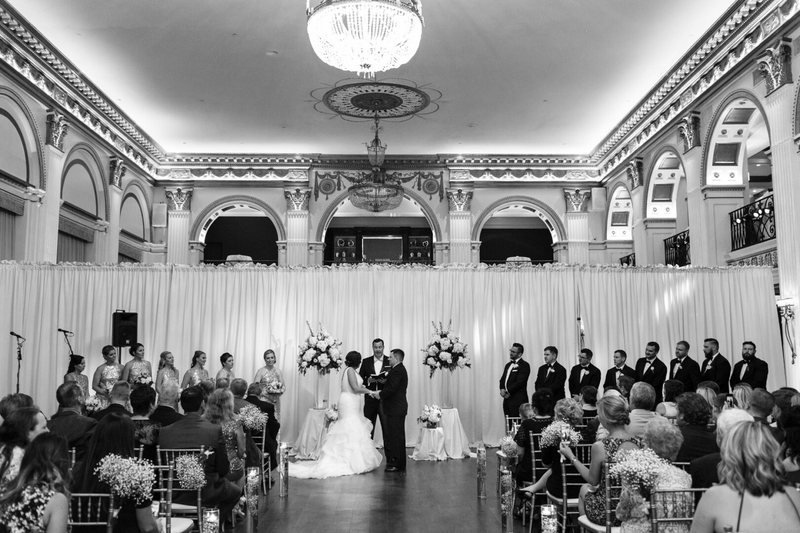 A black-and-white photo captures a Ballroom at the Ben wedding ceremony, with a couple exchanging vows. Bridesmaids and groomsmen stand beside them, while guests are seated in an ornate hall with high ceilings and a chandelier.