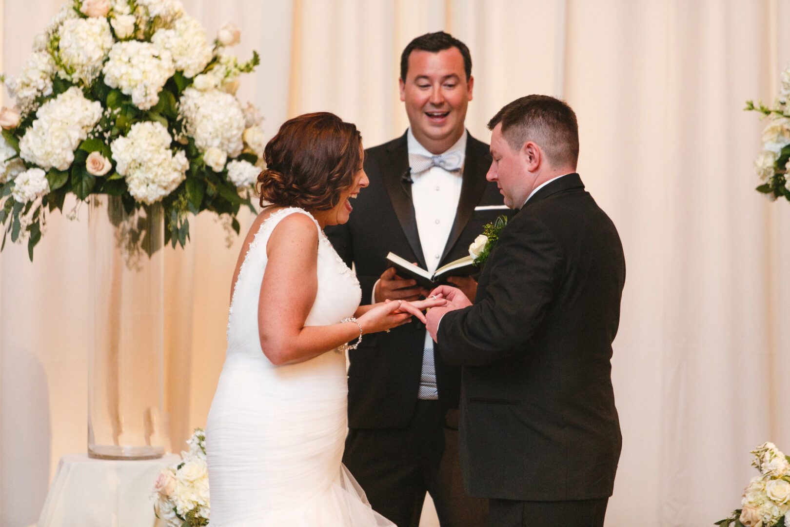 A bride and groom exchange rings during their Ballroom at the Ben wedding ceremony. The officiant stands between them, holding a book, as elegant floral arrangements perfect the scene.