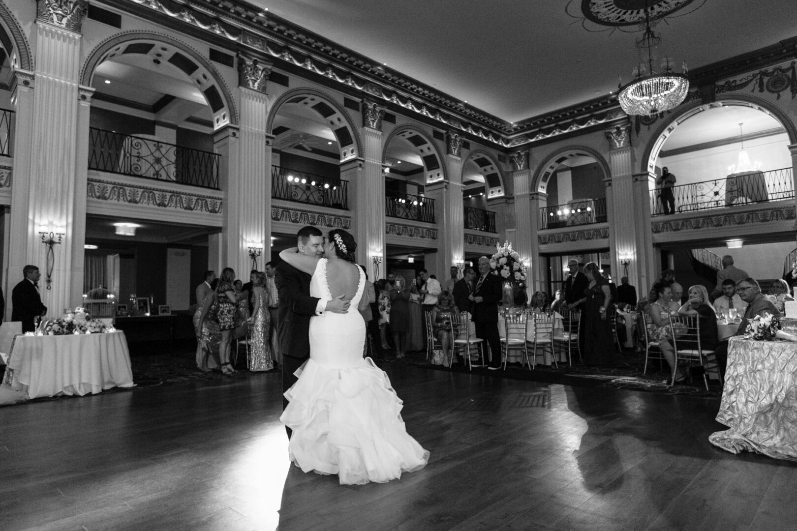 A couple gracefully dances in the grand Ballroom at the Ben, surrounded by guests who are seated and standing. The room's elegant arches and chandeliers add to the enchanting wedding atmosphere.