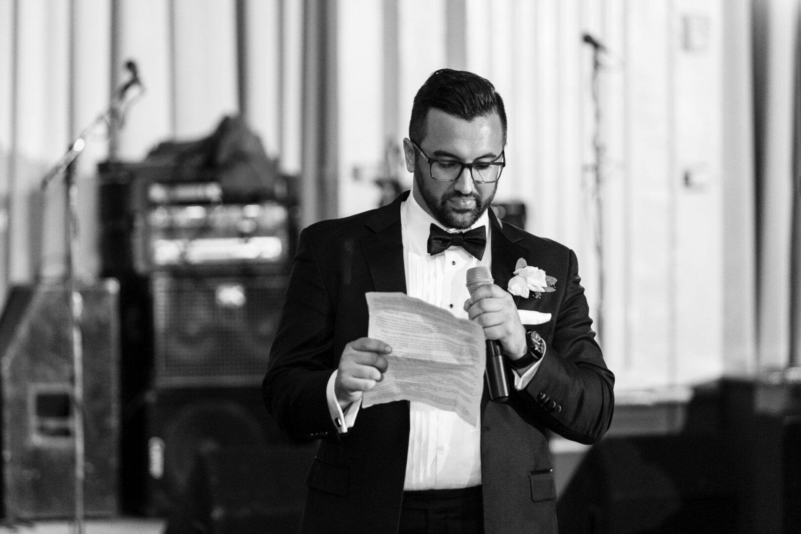 A man in a suit and bow tie stands in the elegant Ballroom at the Ben, reading from a paper while holding a microphone, adding sophistication to the indoor scene.