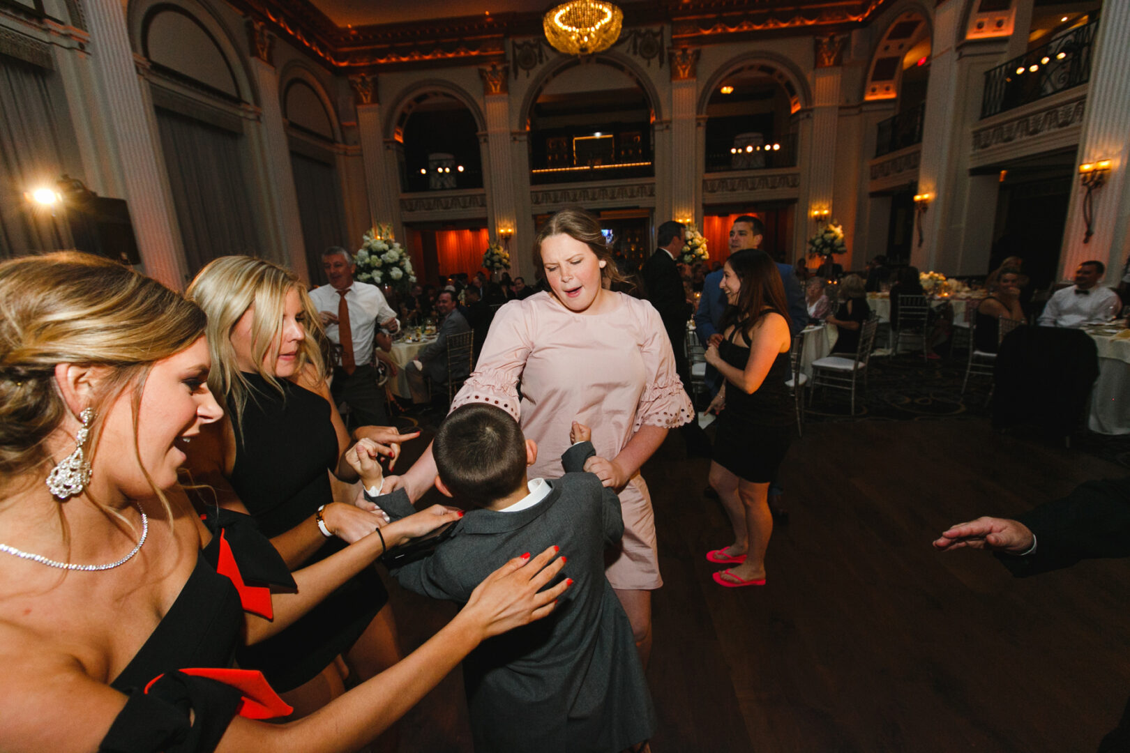 In the magnificent Ballroom at the Ben, people dance energetically among the ornate decor. A woman in a pink dress holds hands with a young boy in a suit, while other guests watch and revel in the lively wedding atmosphere.