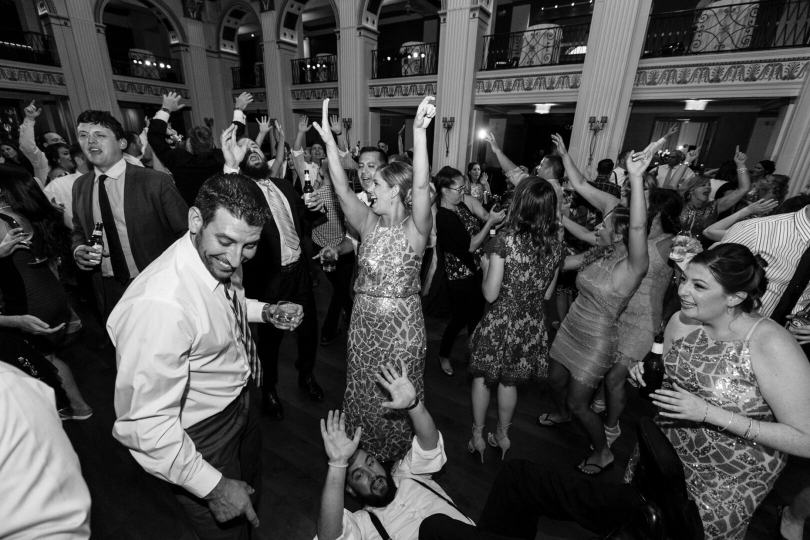A lively crowd in formal attire dances energetically at the Ballroom at the Ben wedding. Amidst the ornate architecture, a man sits on the floor with his arms raised, adding to the joyful scene.
