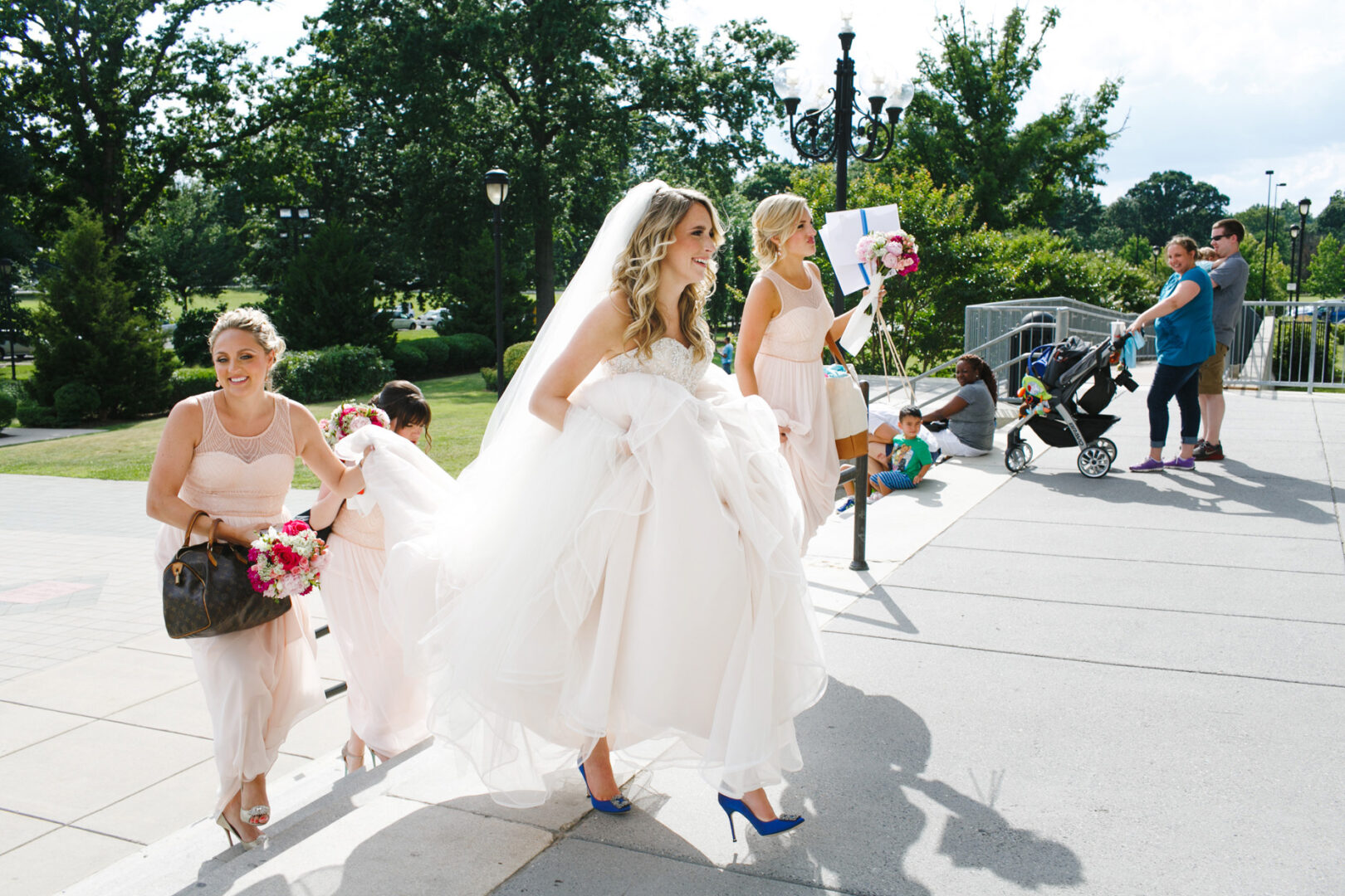 A bride in a white gown and blue heels walks outdoors with bridesmaids at the Please Touch Museum. People with children are nearby. Trees and a sunny sky enhance this joyful wedding scene.