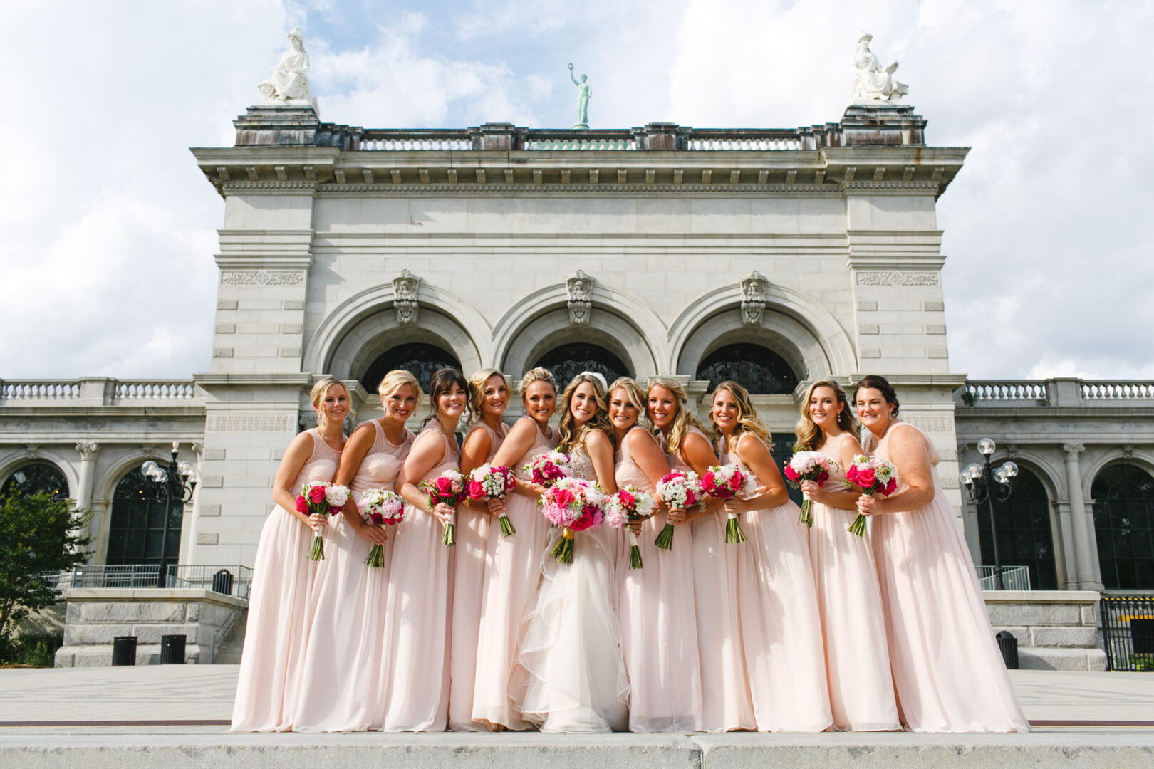 A group of eleven individuals in pink dresses stands in a line, holding bouquets, in front of the grand arches of the Please Touch Museum.