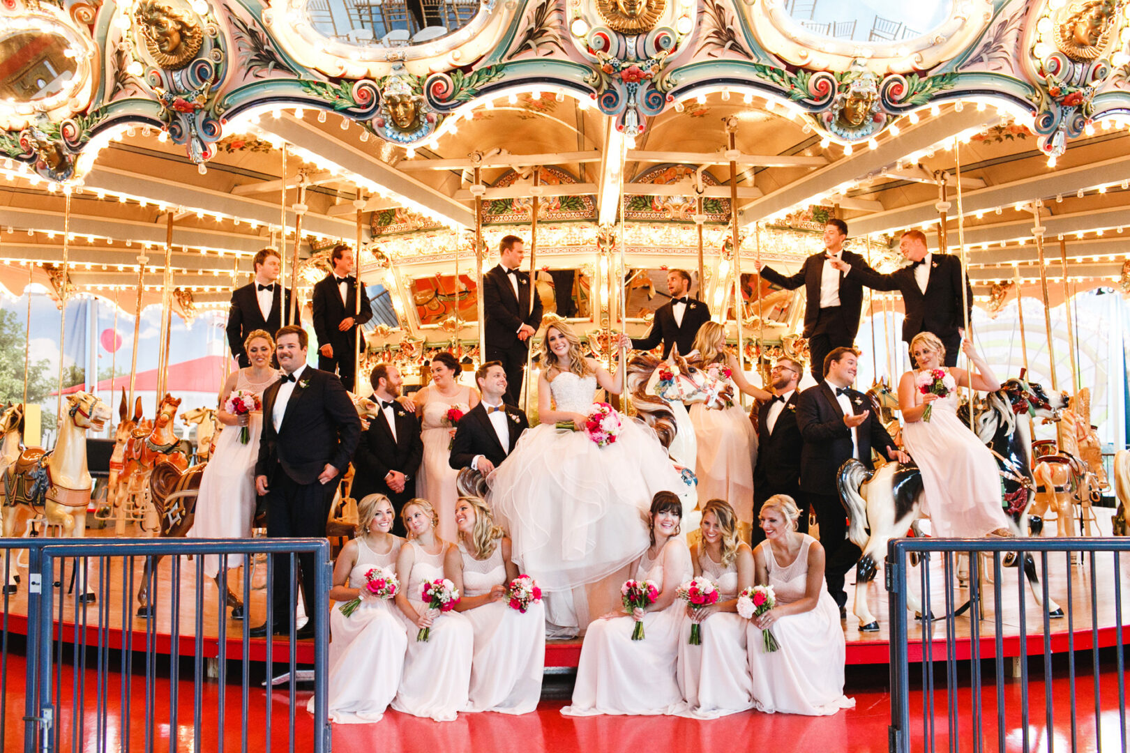 The vibrant wedding party posed joyfully on a carousel at the Please Touch Museum, with the bride, groom, and bridesmaids in white dresses holding pink bouquets, accompanied by groomsmen in sleek black suits.