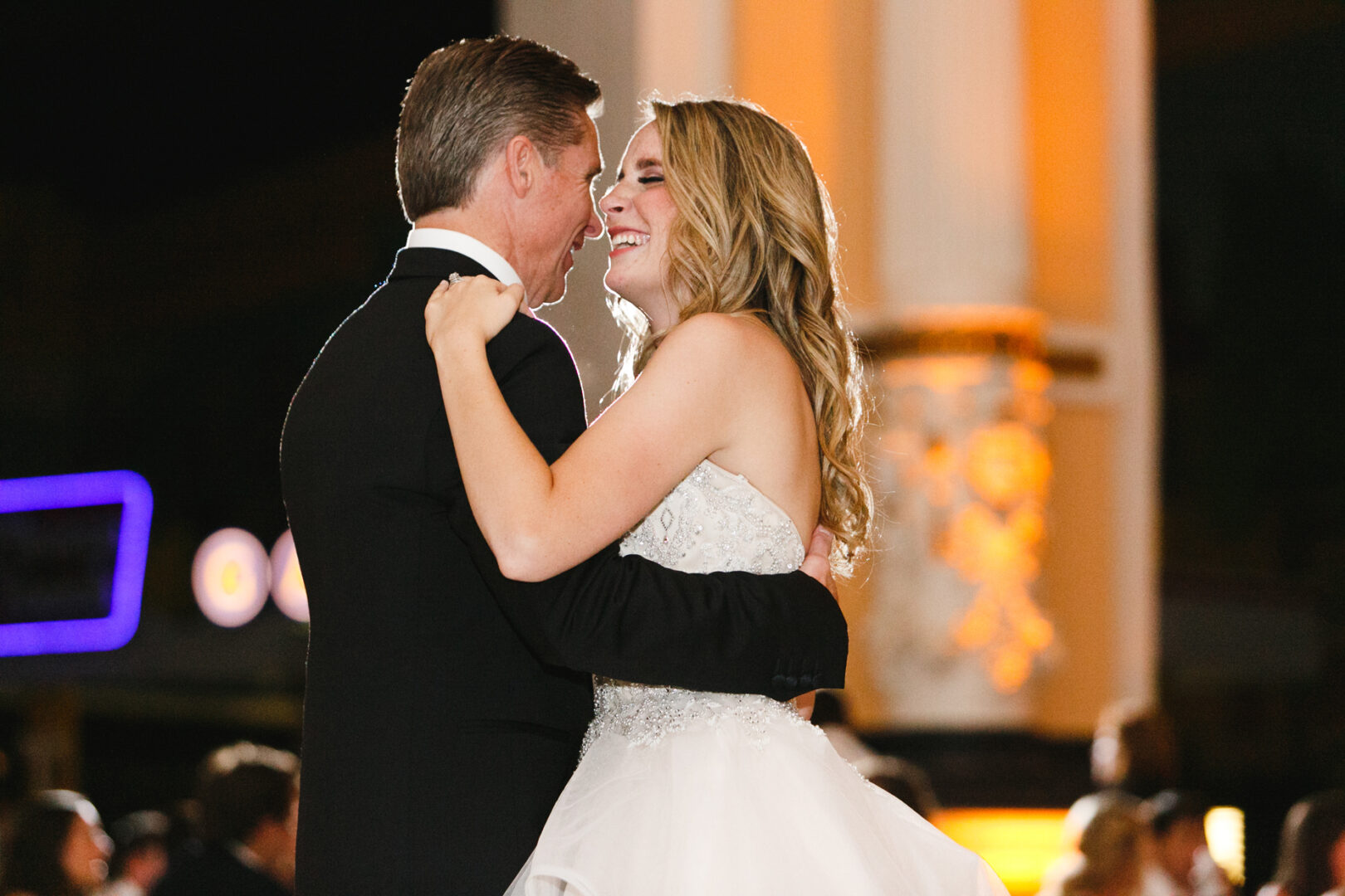 A bride and groom smile and embrace while dancing at a Please Touch Museum wedding reception, illuminated by warm lights in the background.