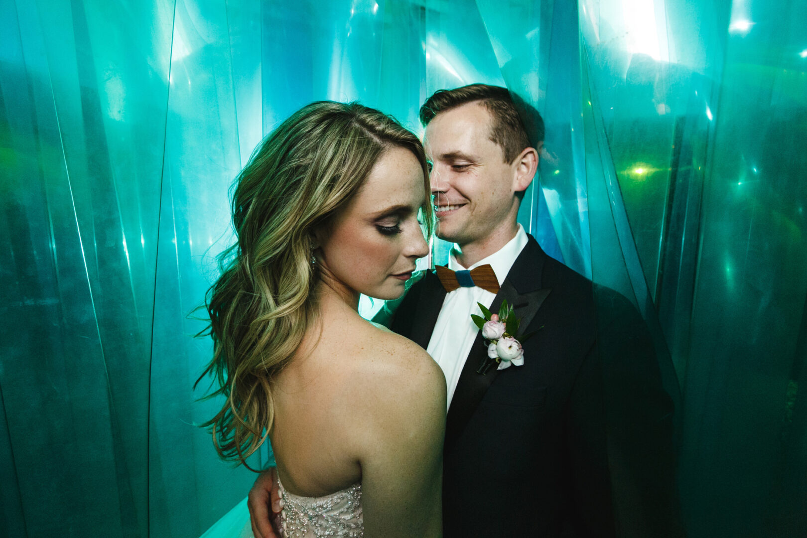 A couple in formal attire stands close, the woman facing left as the man smiles at her amidst a dreamy, teal translucent curtain backdrop, evoking the whimsical charm of a Please Touch Museum wedding.