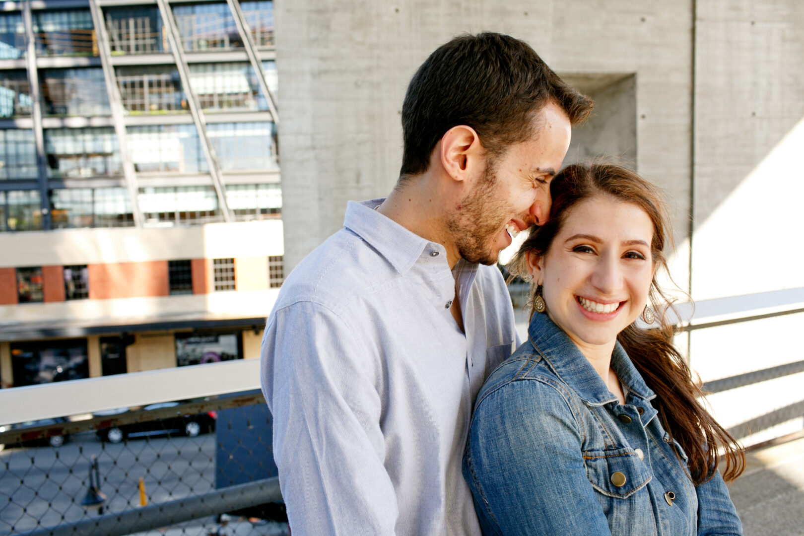 A man and woman stand closely on an urban balcony, smiling during their New York City engagement session. He sports a light blue shirt, while she dons a denim jacket, with blurred city buildings setting the perfect backdrop for this special moment.