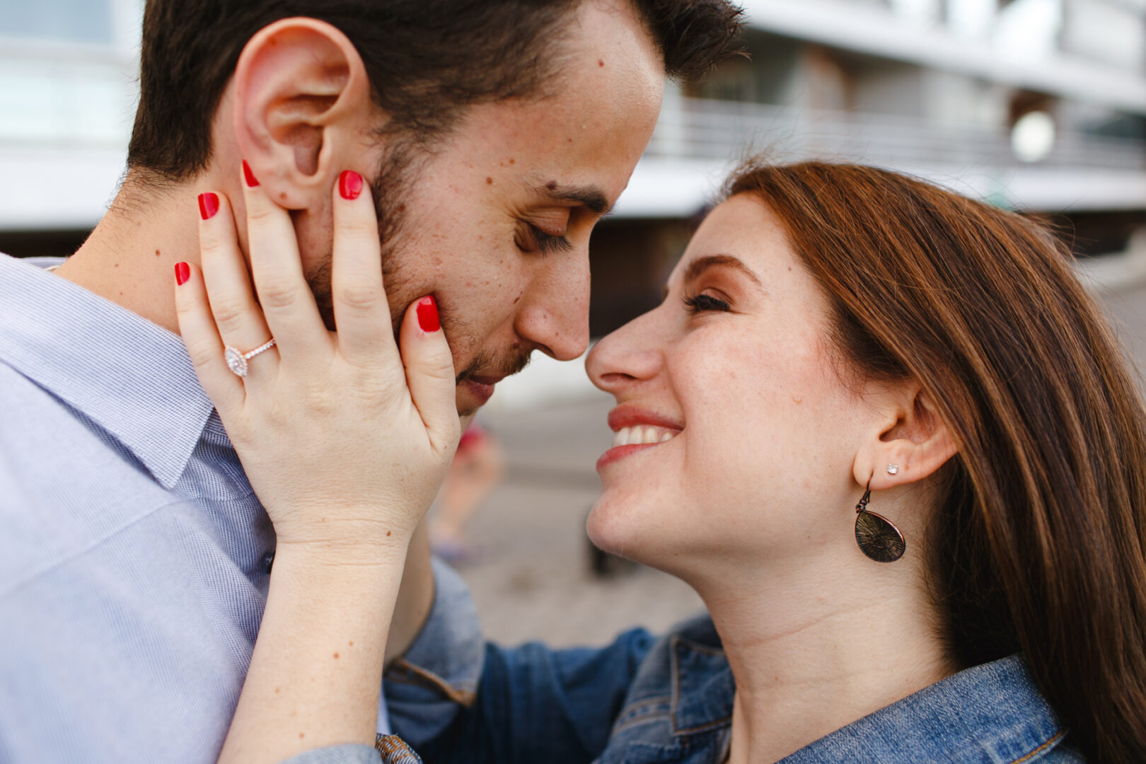 During a New York City engagement session, a woman with red nail polish smiles while holding the face of her bearded partner. They are close together, and she wears a ring on her left hand, capturing a moment of pure joy in the bustling city.