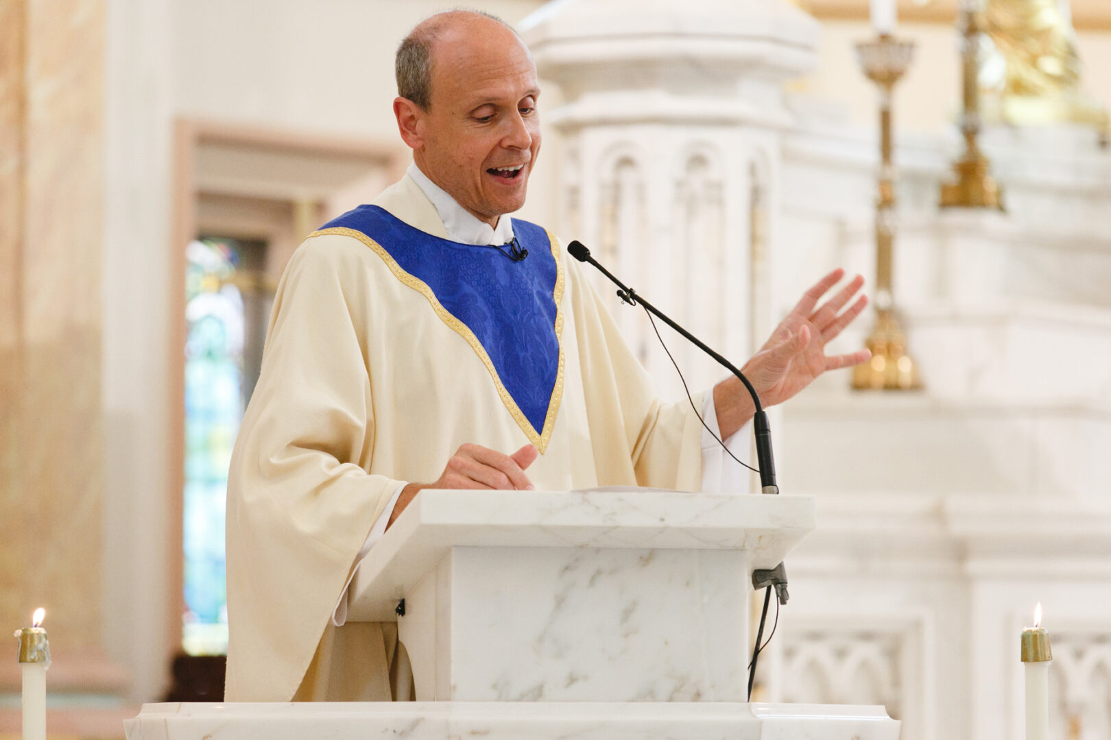 A man in a church robe speaks at a marble lectern, holding a microphone. Candles are visible in the foreground, casting a warm glow reminiscent of an intimate Congress Hall wedding ceremony.