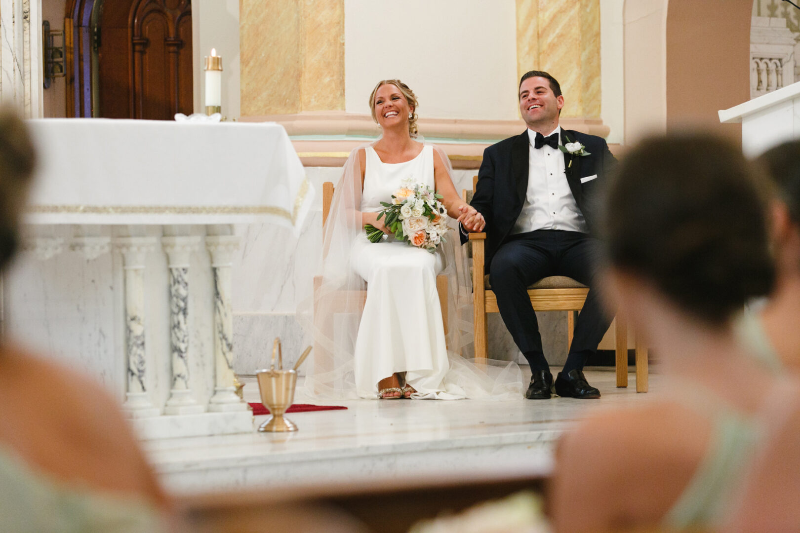 A bride and groom sit smiling in a church at their Congress Hall wedding, holding hands. A bouquet rests on the bride's lap, while bridesmaids can be seen from behind in the foreground.