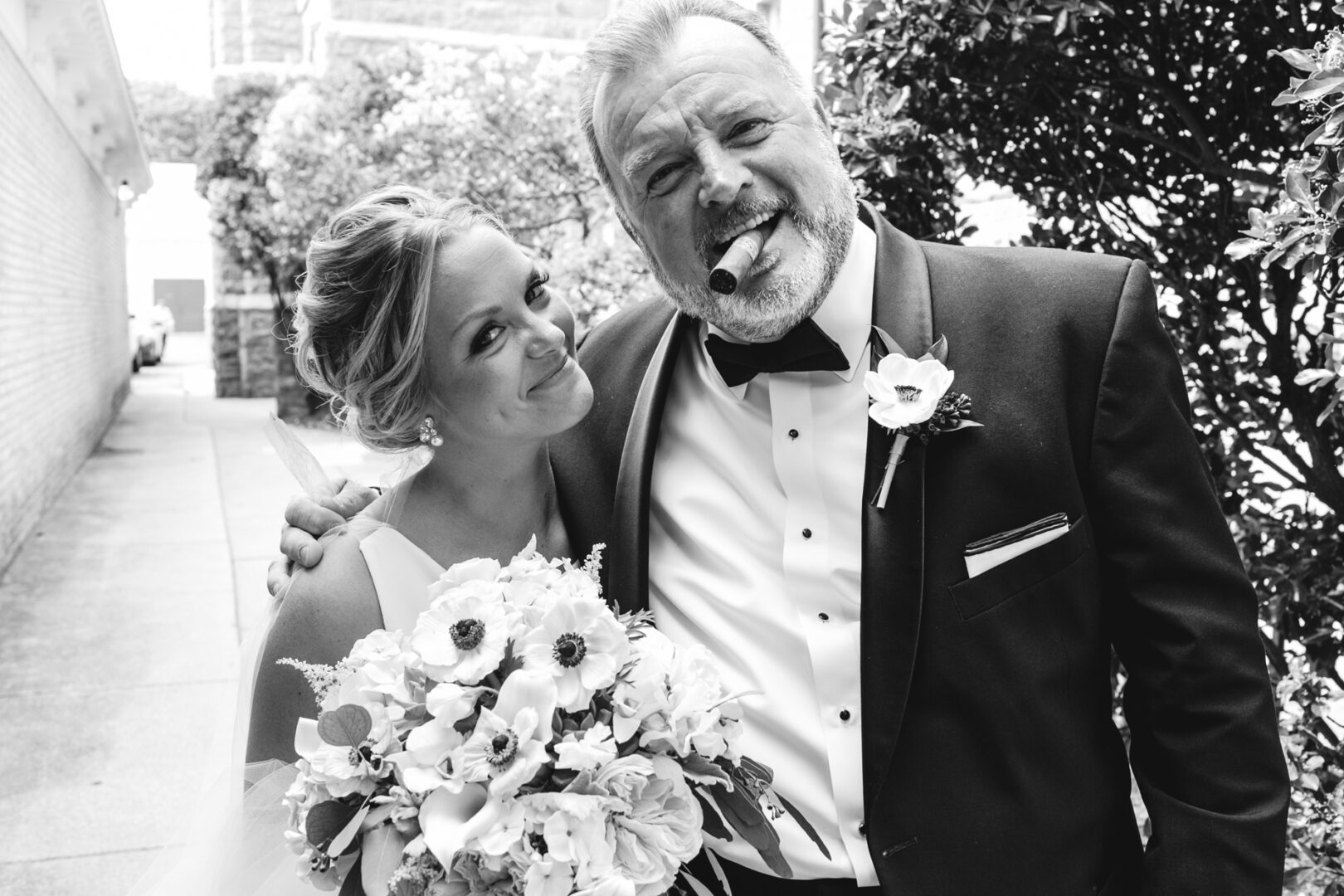 Bride and older man in formal attire smiling outdoors at a Congress Hall wedding; the bride holds a bouquet, while the man enjoys a cigar.