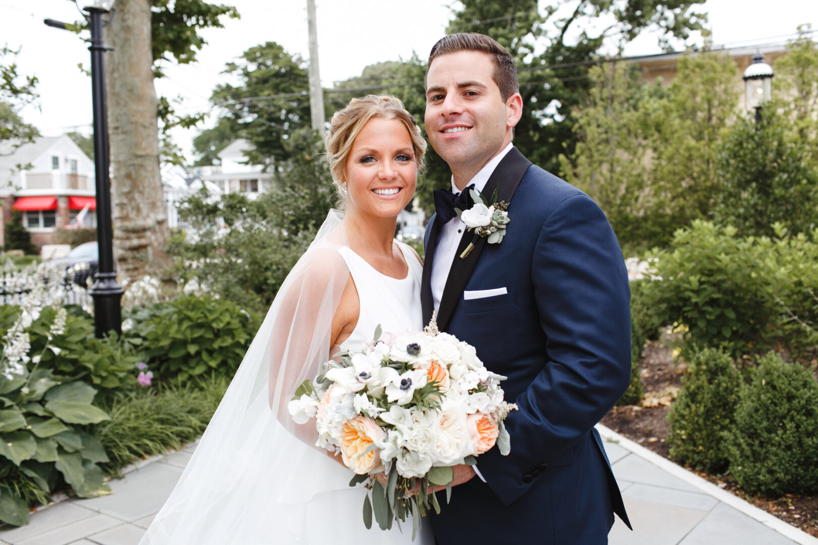 The bride and groom beam with joy outdoors at their Congress Hall wedding, as the bride holds a white and peach bouquet amidst lush greenery.