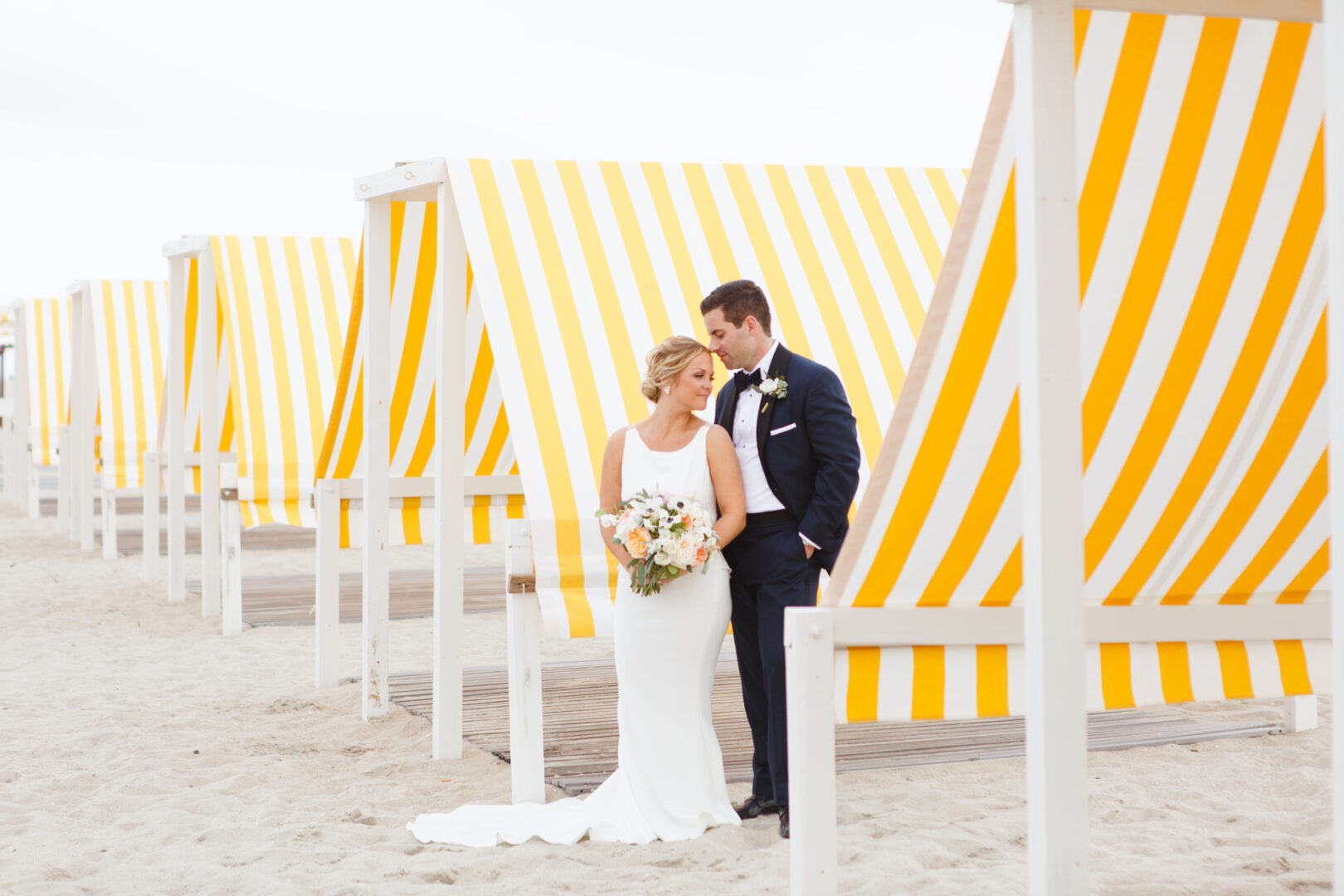 The bride and groom stand together on a sandy beach, in front of yellow and white striped cabanas at their enchanting Congress Hall wedding. The bride holds a bouquet.