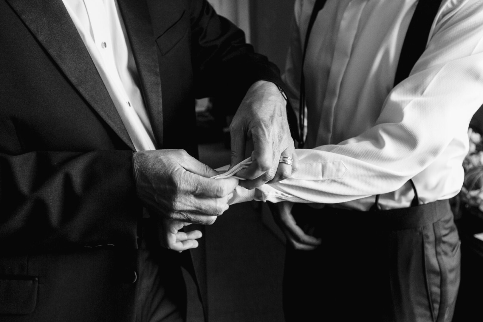 Two individuals adjusting cufflinks on a shirt sleeve; one in a suit jacket, the other in a dress shirt with suspenders. This classic black and white image captures the timeless elegance of preparations at a Glen Island Harbour Club wedding.