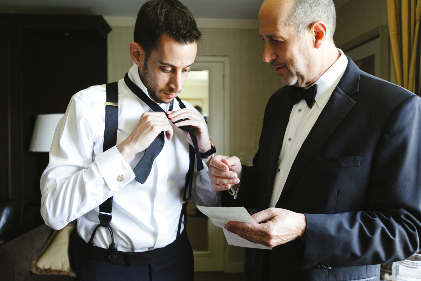 Two men in formal attire, one adjusting his tie while the other holds a paper, stand in a room with a sofa and lamp in the background, preparing for a Glen Island Harbour Club wedding.