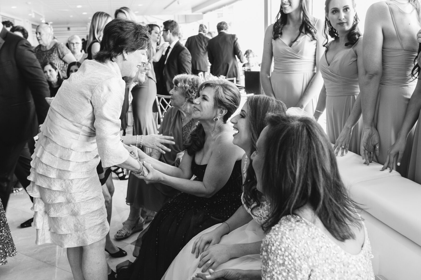 Amidst the elegance of a Glen Island Harbour Club wedding, a seated woman in a black dress greets a standing woman in a light-colored gown as other attendees chat and mingle around them.