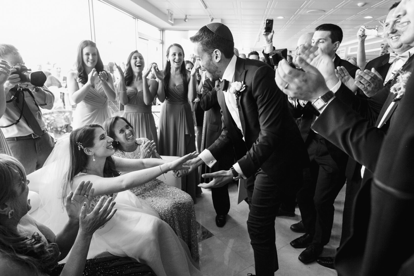 At a Glen Island Harbour Club wedding, a bride and groom smile and hold hands, surrounded by applauding guests in formal attire at the celebration.