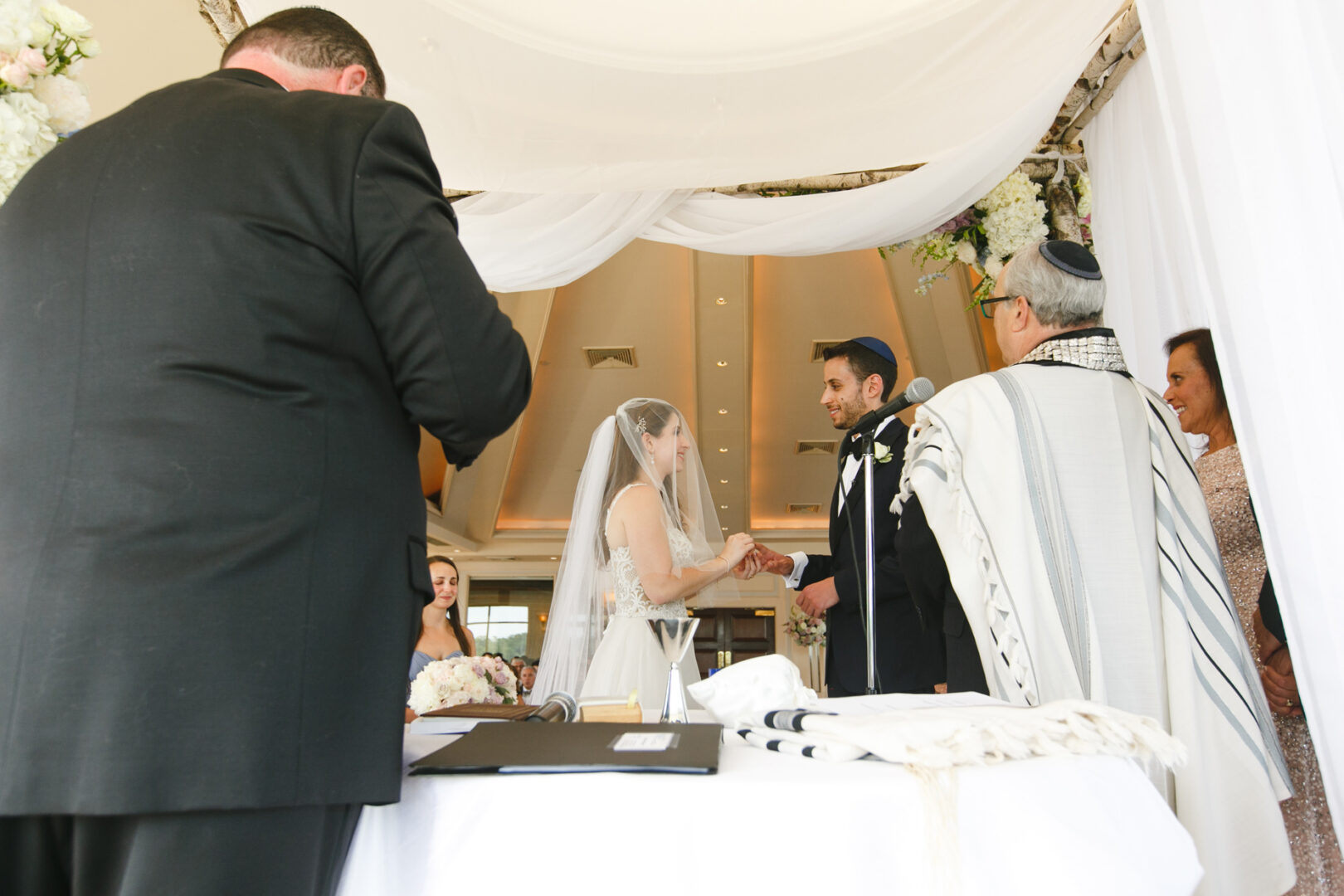 During their Glen Island Harbour Club wedding, a bride and groom exchange rings under a white canopy. With the photographer capturing every moment, the officiant guides the ceremony while guests look on, surrounded by the scenic beauty of the harbor.