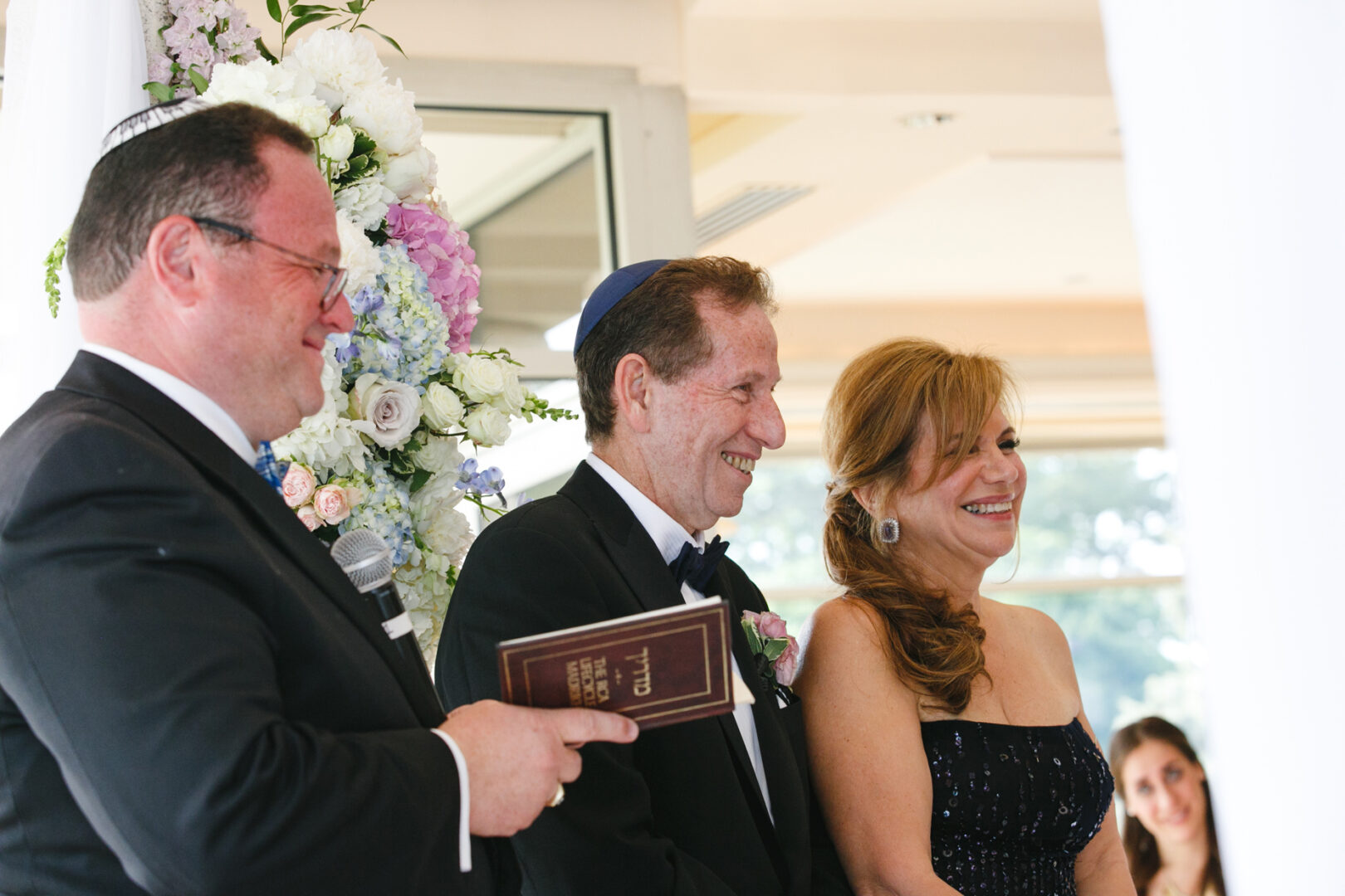 Three people stand together at a Glen Island Harbour Club wedding. One holds a microphone and a book. Elegant floral arrangements provide the perfect backdrop for this formal event.