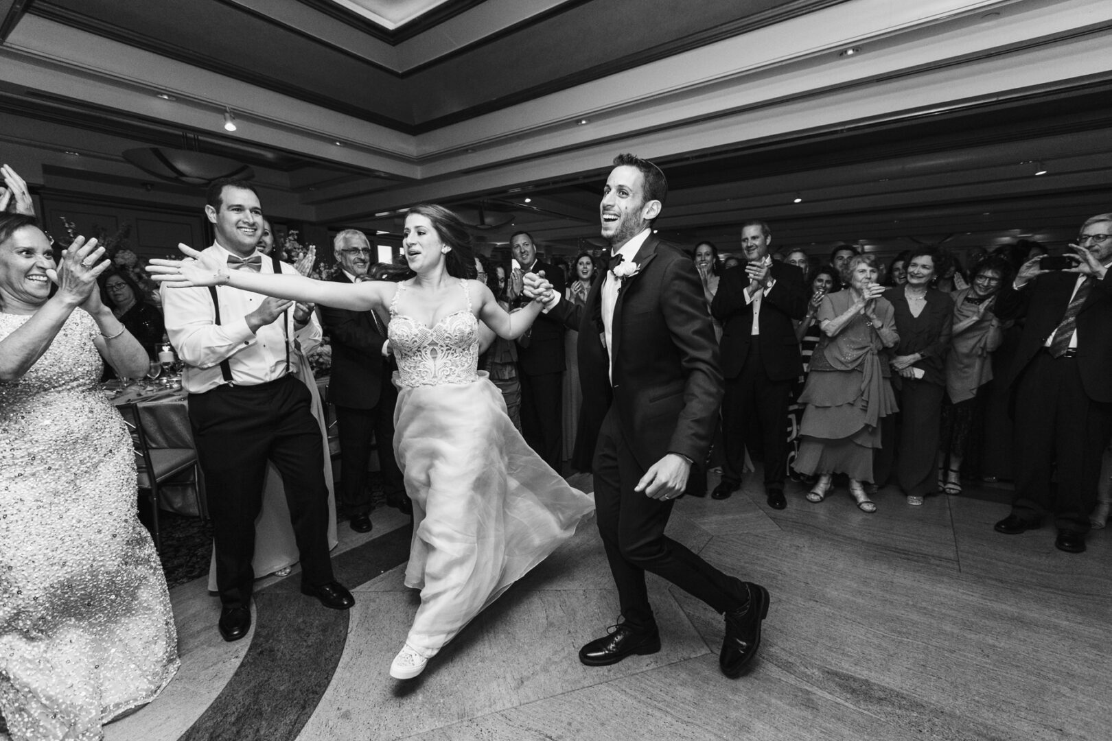The bride and groom dance joyfully in a crowded reception hall at Glen Island Harbour Club, surrounded by clapping guests.