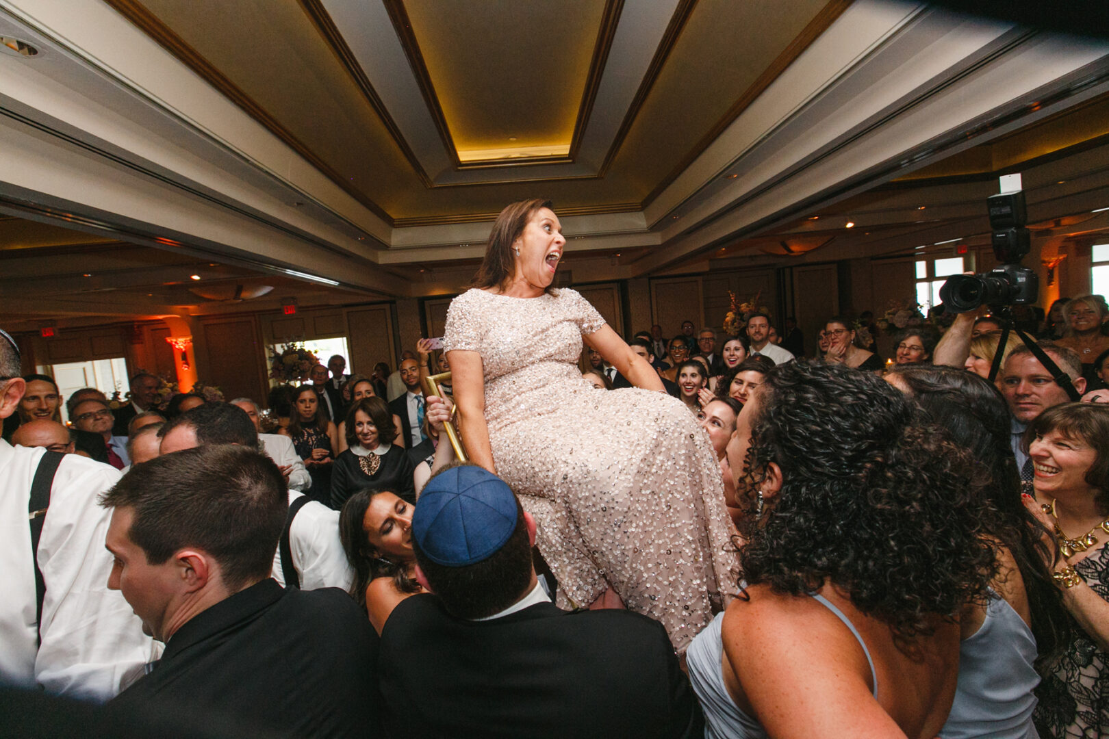 At a Glen Island Harbour Club wedding, a woman in a light-colored dress is lifted on a chair by joyful guests during the celebration.