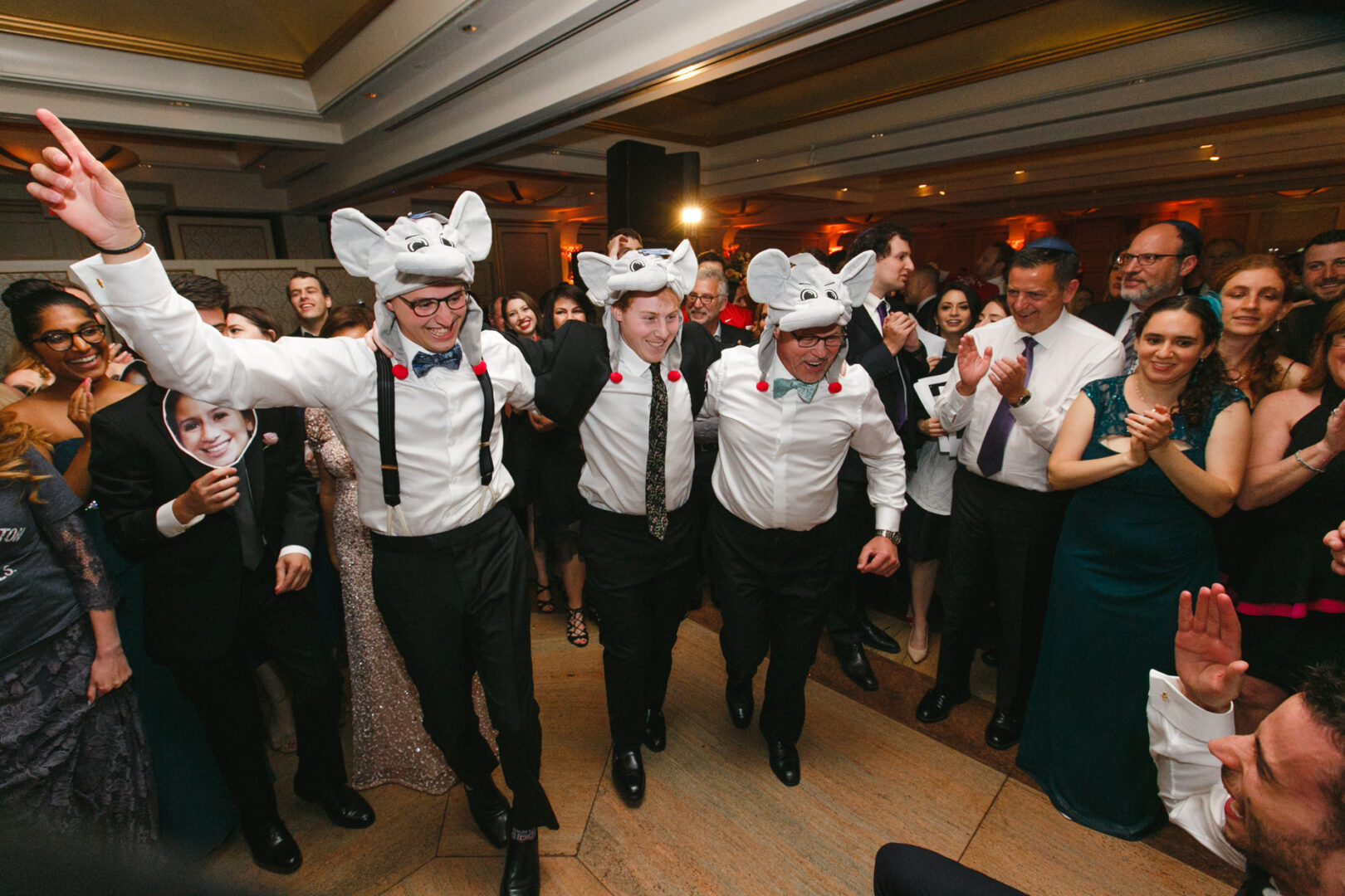 Three men wearing matching hats and shirts dance joyfully at a Glen Island Harbour Club wedding, surrounded by a crowd of applauding guests.