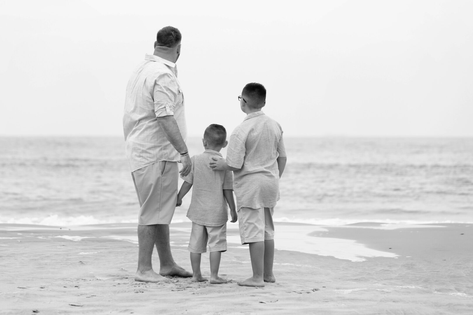 A man and two boys stand on the beach, facing the ocean in a classic Ocean City family photo. They are barefoot and wearing similar casual outfits, perfectly capturing a timeless moment. The scene is in black and white, adding a nostalgic touch to their shared adventure.