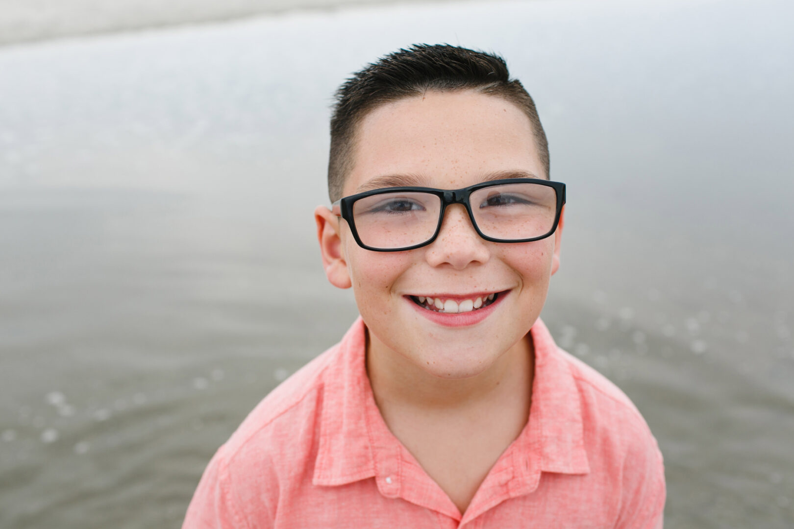 A boy with short hair and glasses smiles, wearing a pink shirt, standing near the shimmering waters of Ocean City.