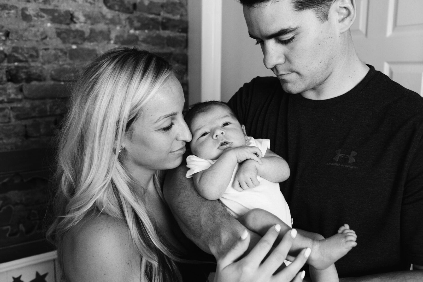 A couple holds a baby, with the woman gently touching the tiny hand. They stand indoors against a timeless brick wall backdrop, capturing the warmth of Philadelphia newborn photos.