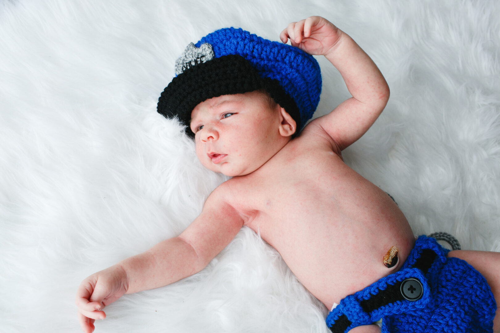 A baby rests on a white fuzzy surface, donning a crocheted blue and black police-themed outfit—a perfect capture for Philadelphia newborn photos.