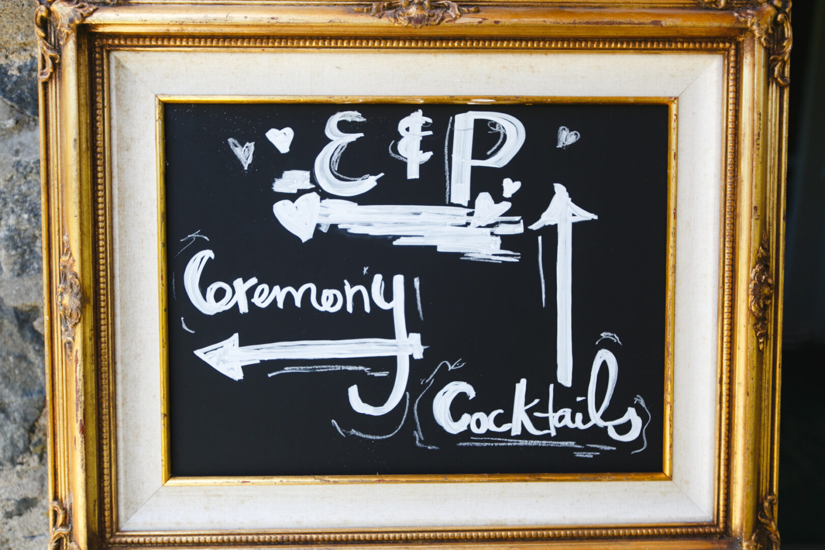 Chalkboard sign in a gold frame with arrows and hearts, directing guests at the Rockwood Park Carriage House Wedding to "Ceremony" on the left and "Cocktails" on the right. Initials "E & P" grace the top.