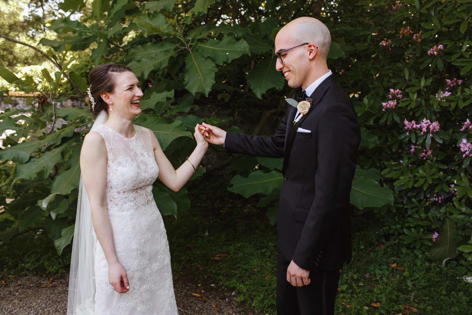 A bride and groom smile at each other, holding hands amidst the lush greenery of Rockwood Park, their wedding unfolding enchantingly by the charming Carriage House.