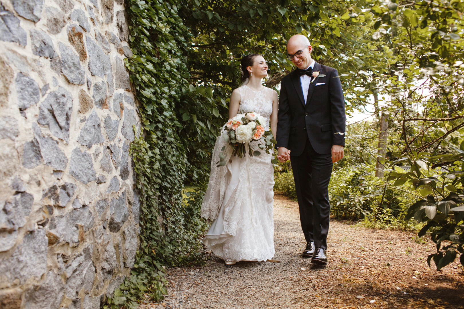 At their Rockwood Park Carriage House wedding, the bride and groom in formal attire walk hand in hand along a stone wall, surrounded by lush greenery. The bride holds a beautiful bouquet of flowers.