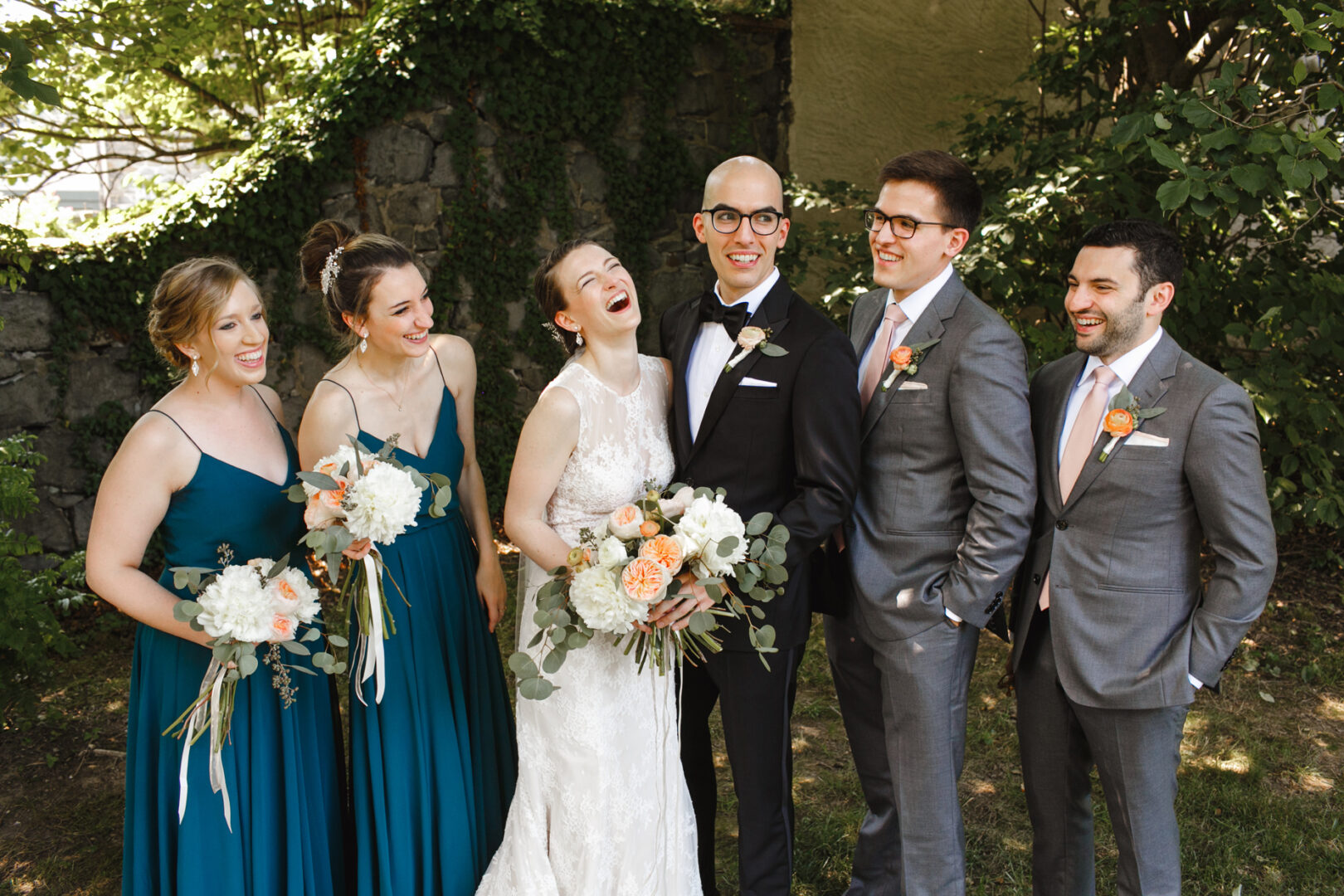 At their Rockwood Park Carriage House wedding, the bride and groom stand laughing with their bridesmaids and groomsmen, all dressed formally. The bridesmaids hold bouquets with white and peach flowers.