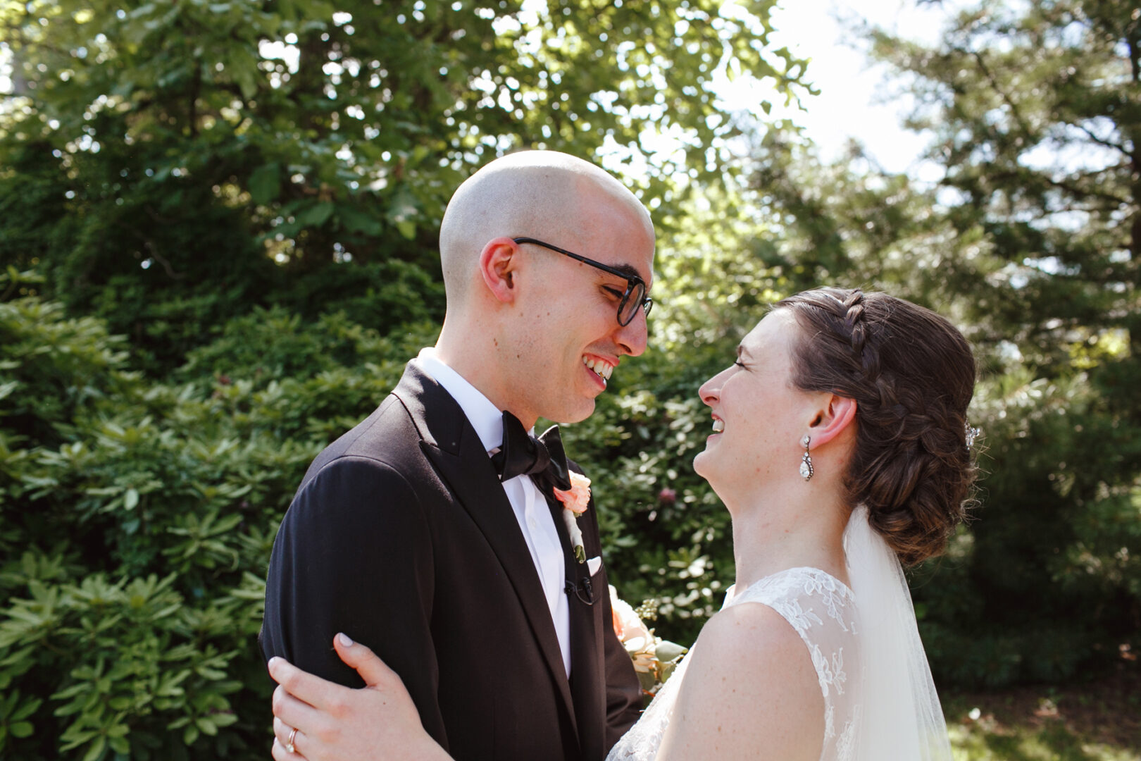 A smiling couple in formal attire stands outdoors at Rockwood Park Carriage House, facing each other amidst greenery and sunlight.