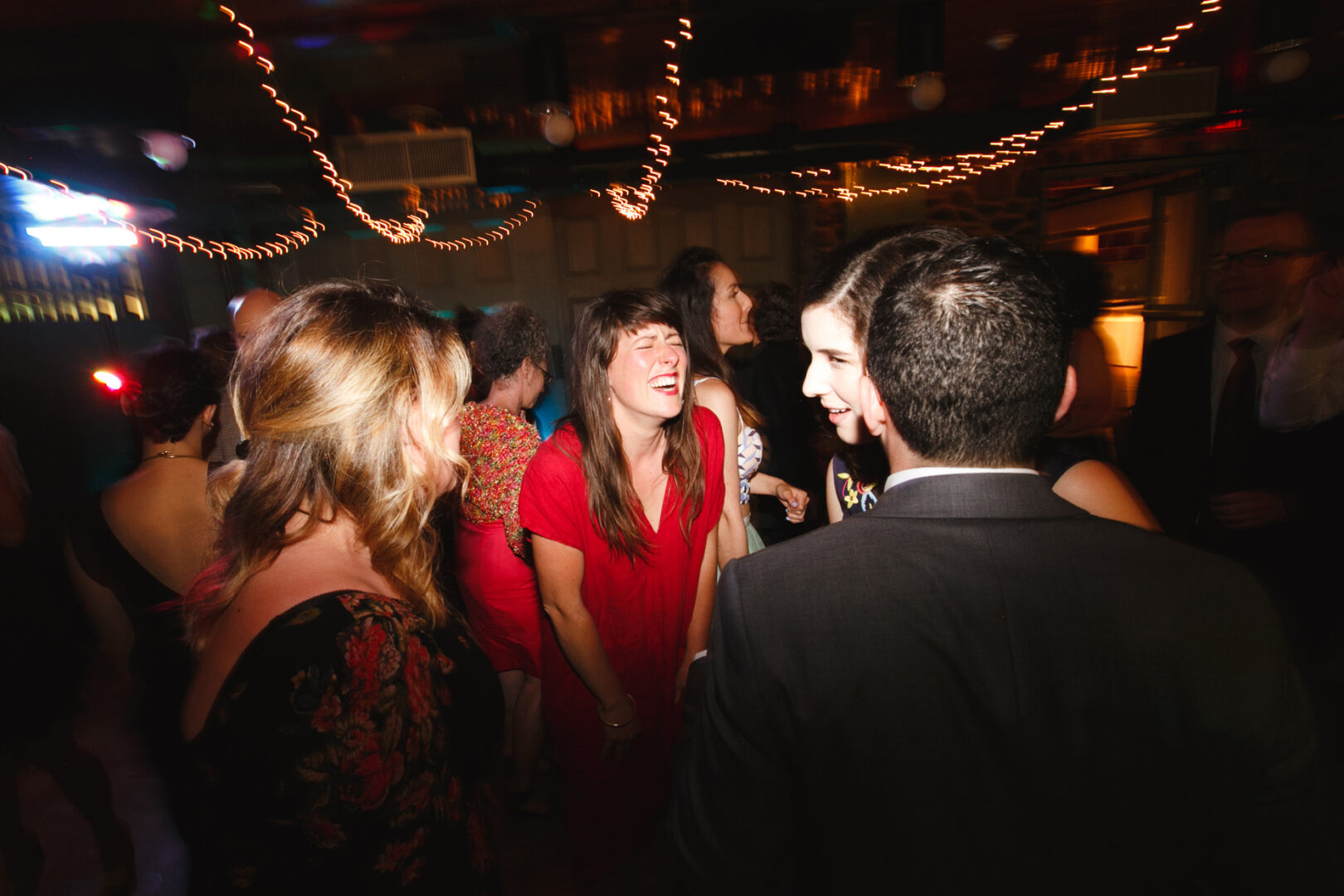 The Rockwood Park Carriage House wedding is in full swing, with people dancing and socializing. The room glows with string lights, and guests in formal attire add a touch of elegance to the celebration.