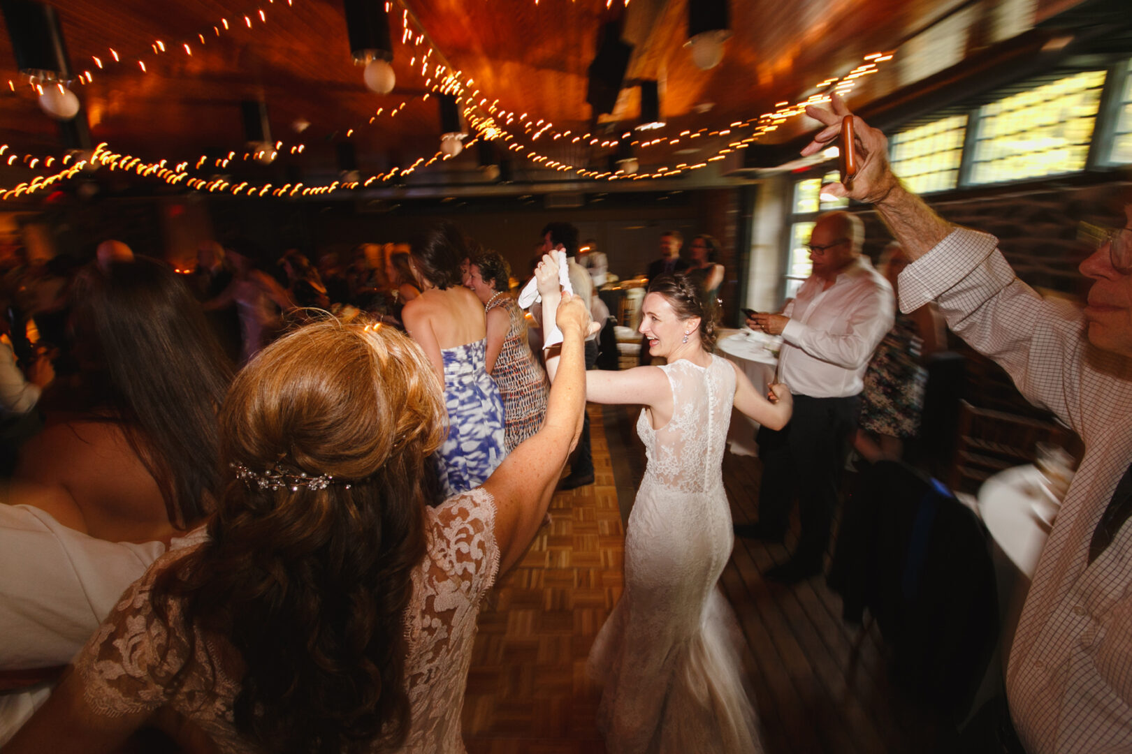 Guests dance energetically in a warmly lit venue at the Rockwood Park Carriage House Wedding, with a bride in a white gown leading the way. String lights hang above, creating a festive atmosphere.