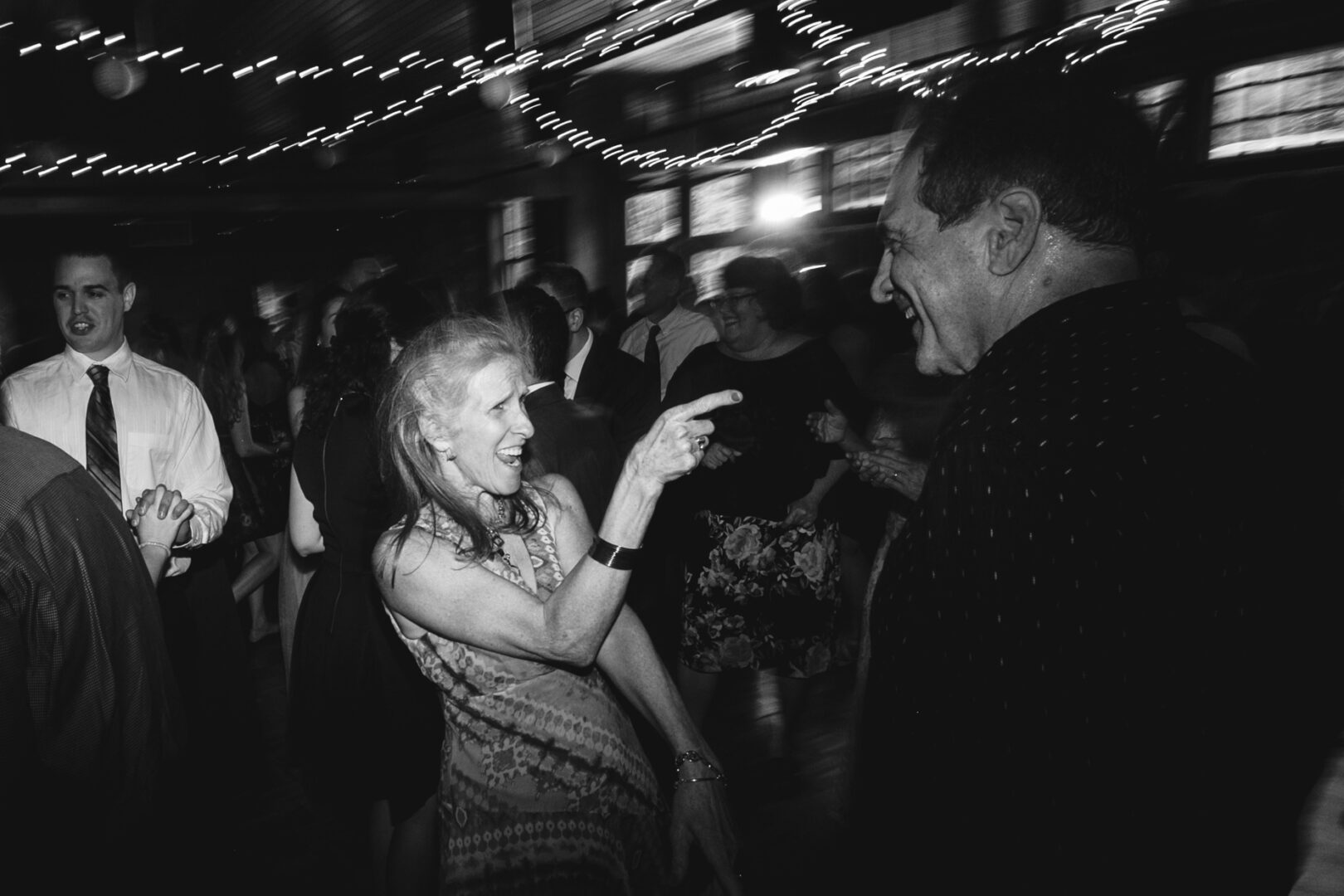 In the enchanting glow of string lights at a Rockwood Park Carriage House wedding, people dance and laugh. A woman playfully points at a man, both beaming with joy amidst the merry crowd.