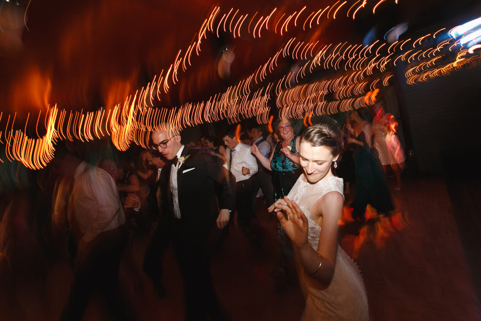 People dancing under string lights at the Rockwood Park Carriage House Wedding, with a blurred motion effect creating dynamic streaks of light.