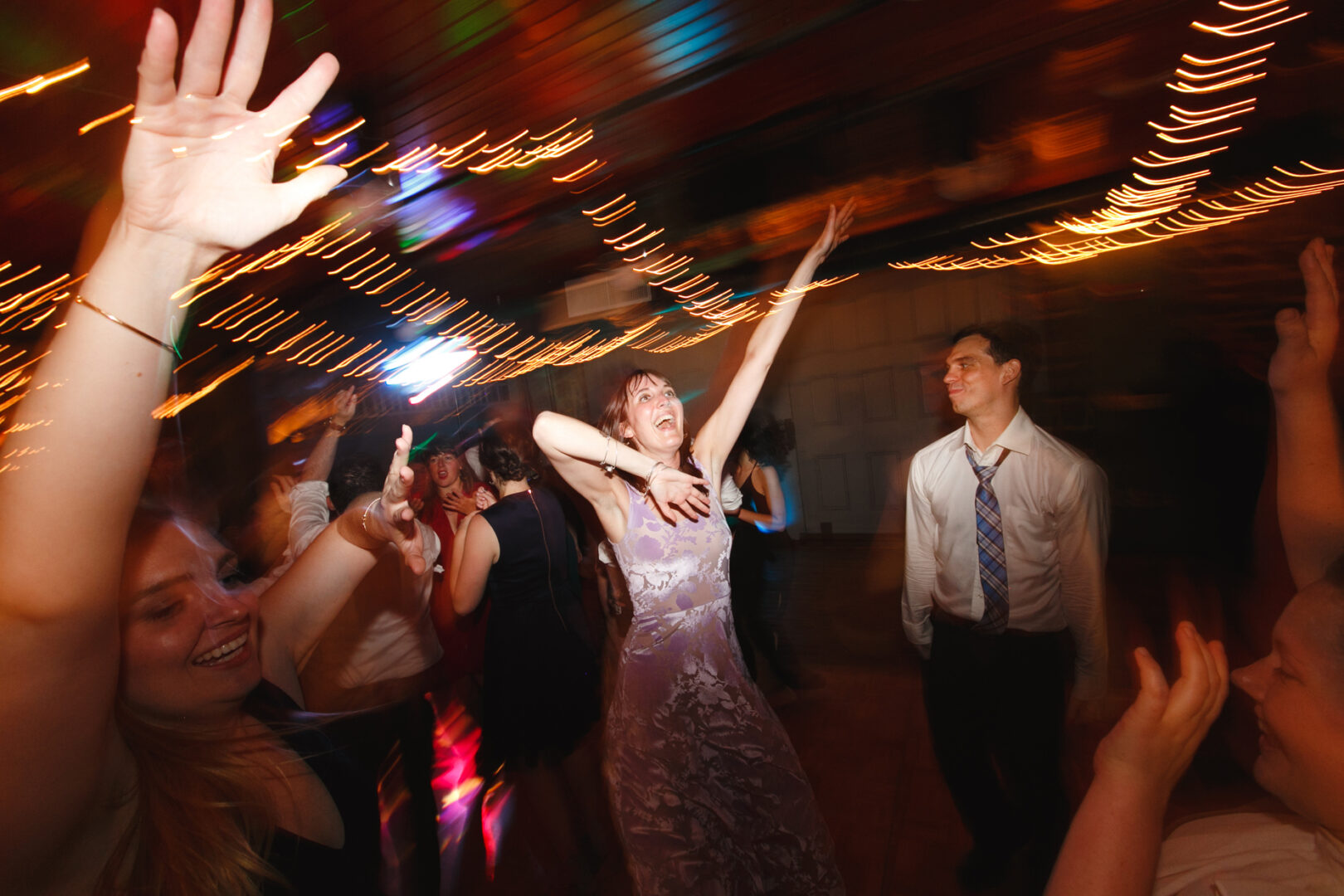 People dancing energetically at a Rockwood Park Carriage House wedding, under colorful string lights, with a woman in the foreground raising her arms.