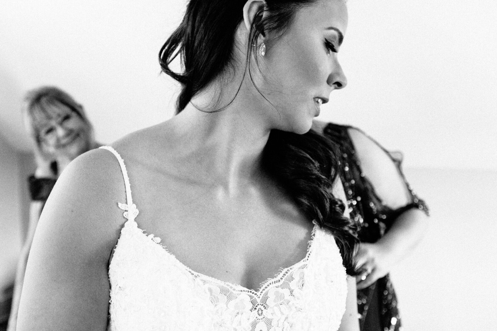 A woman in a white lace dress is captured in profile, embodying the timeless elegance often seen in Barn on Bridge wedding photos. Two people linger in the background of this indoor black and white photograph, adding layers of depth and nostalgia.