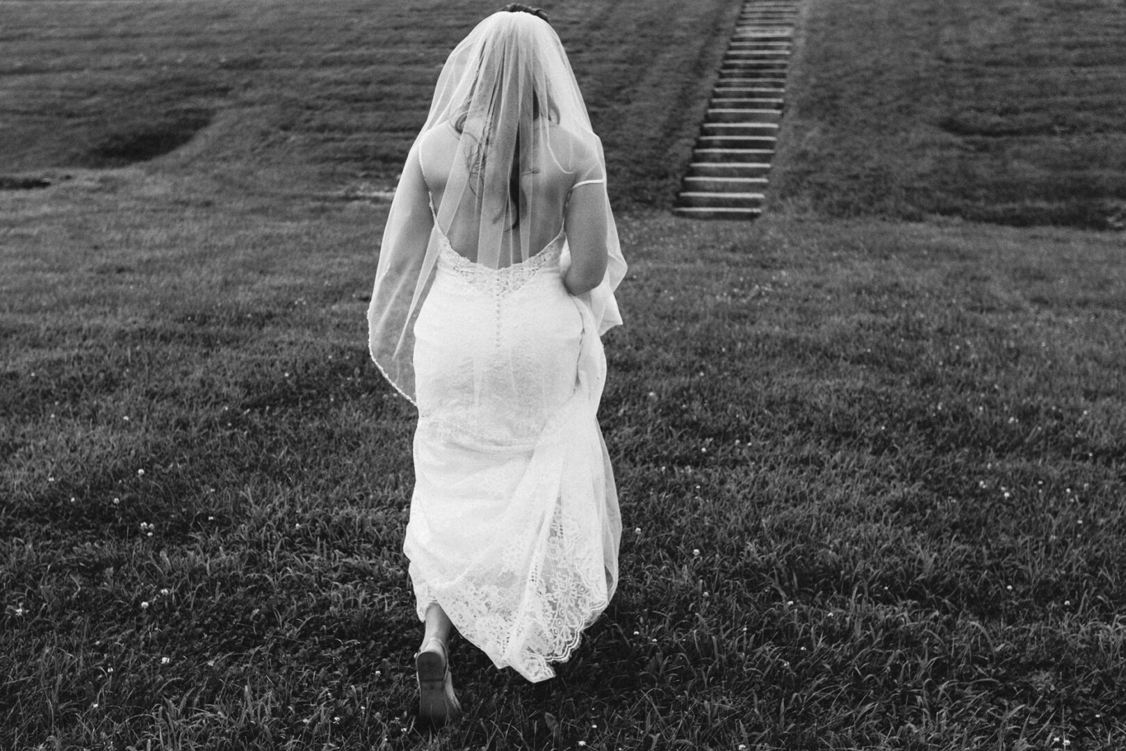 Barefoot on the grass, a bride in a lace gown and veil elegantly approaches a staircase in the field. This enchanting moment captured near the iconic Barn on Bridge makes for timeless wedding photos that tell a magical story.